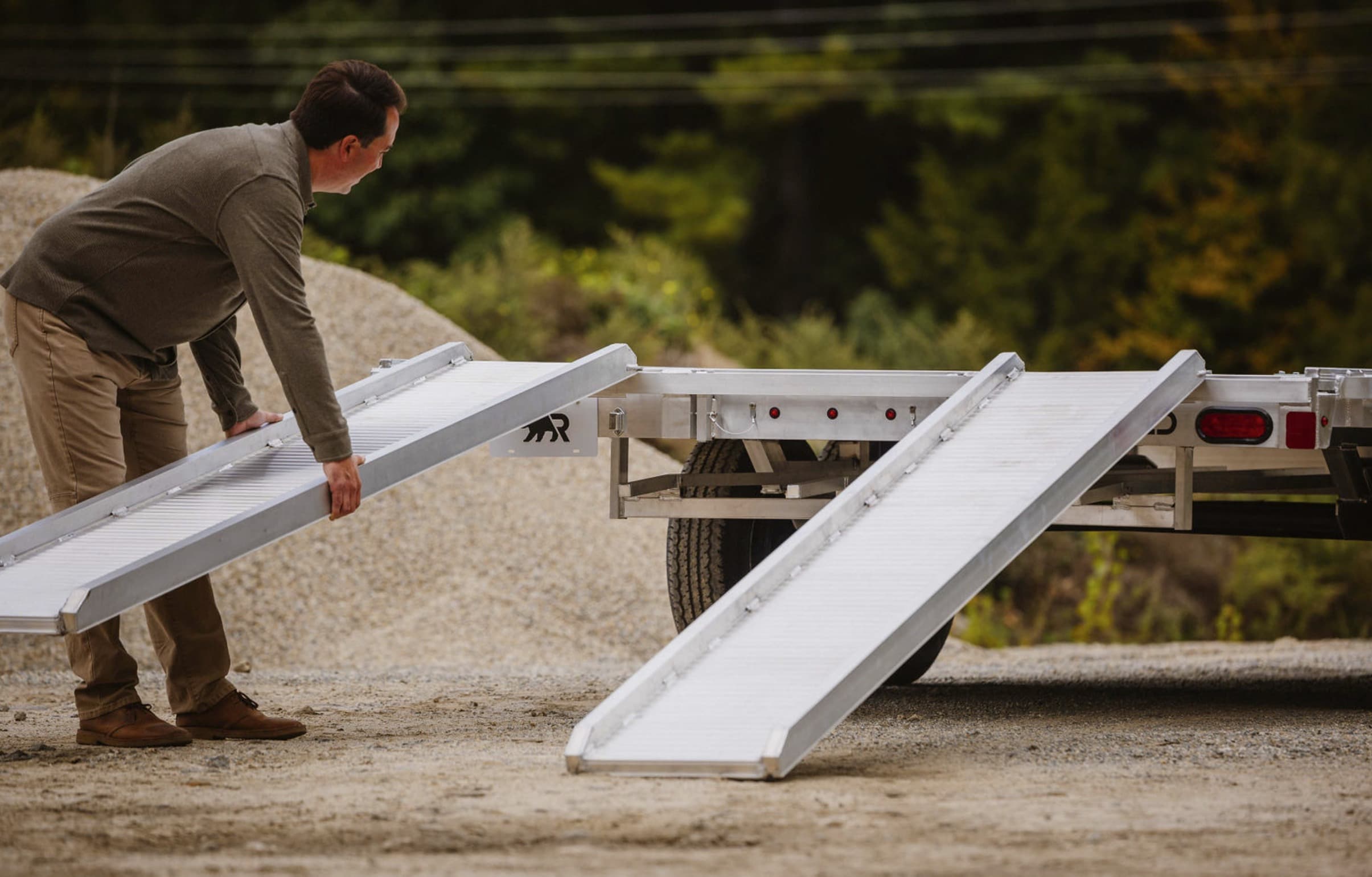 An owner sets an aluminum stow-away ramp onto the rear of a Rugged deckover trailer, with the second ramp already in place.