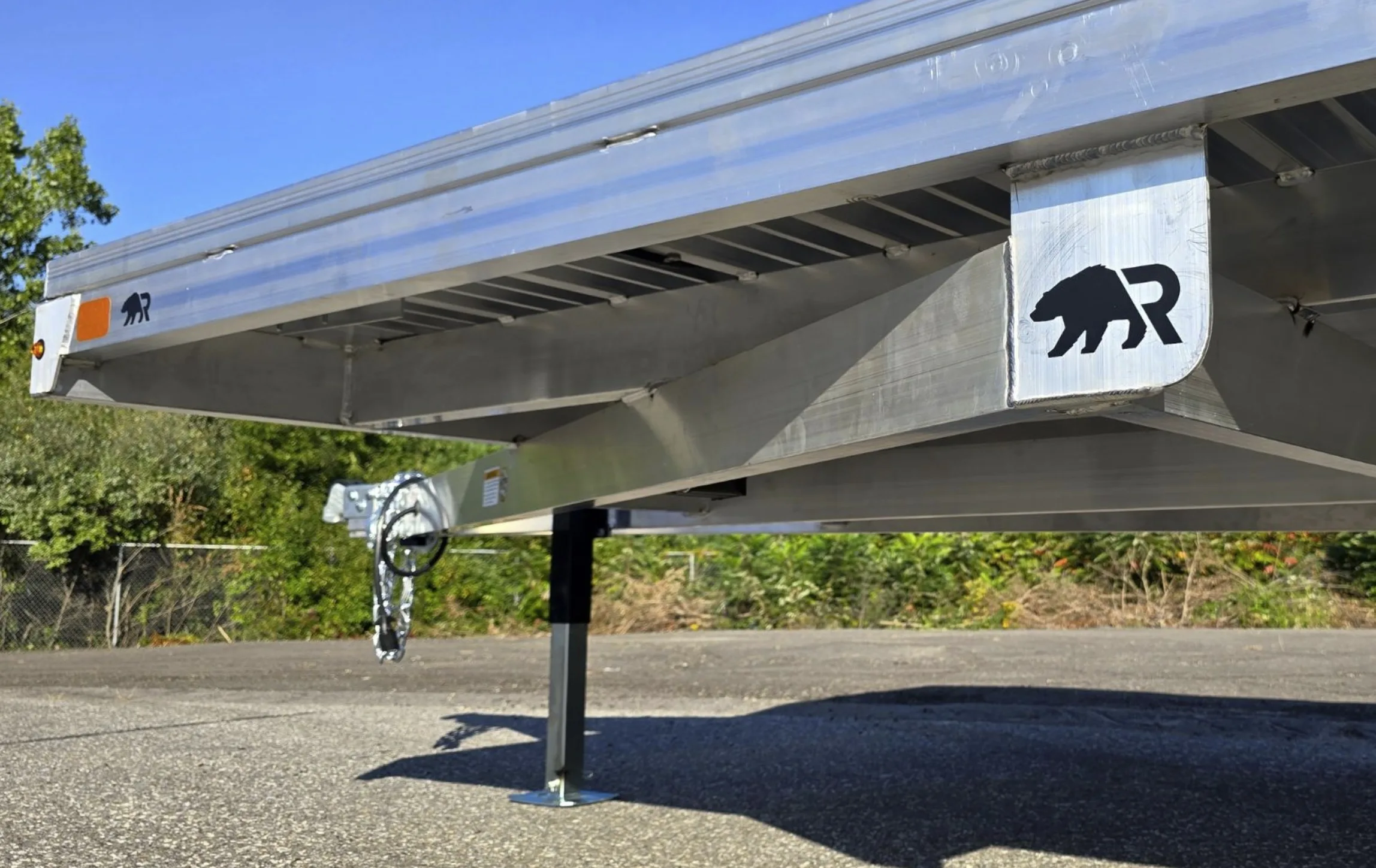 Front underside of a Rugged deckover trailer showing the welded aluminum frame, coupler with safety chains, and drop-leg jack