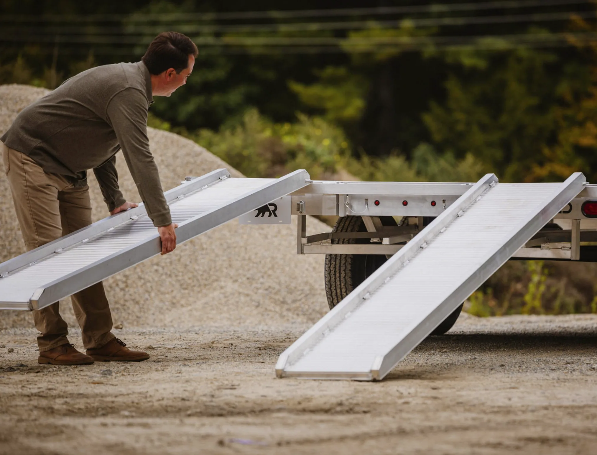 A man positions an aluminum loading ramp against the flat, full-width deck of a Rugged deckover trailer