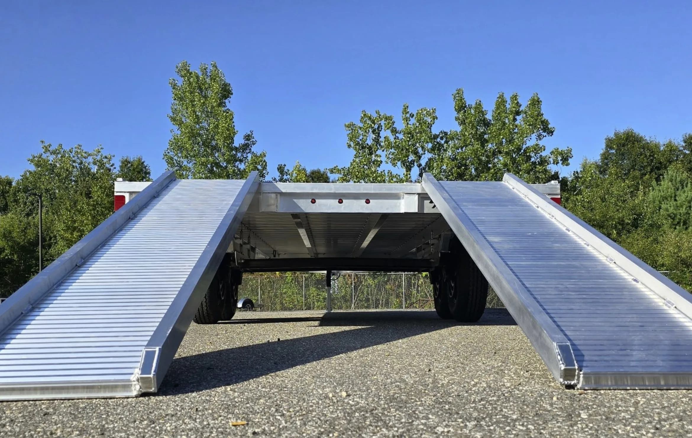 Rear view beneath a Rugged deckover trailer with both ramps down, showing the aluminum deck structure above the tandem wheels