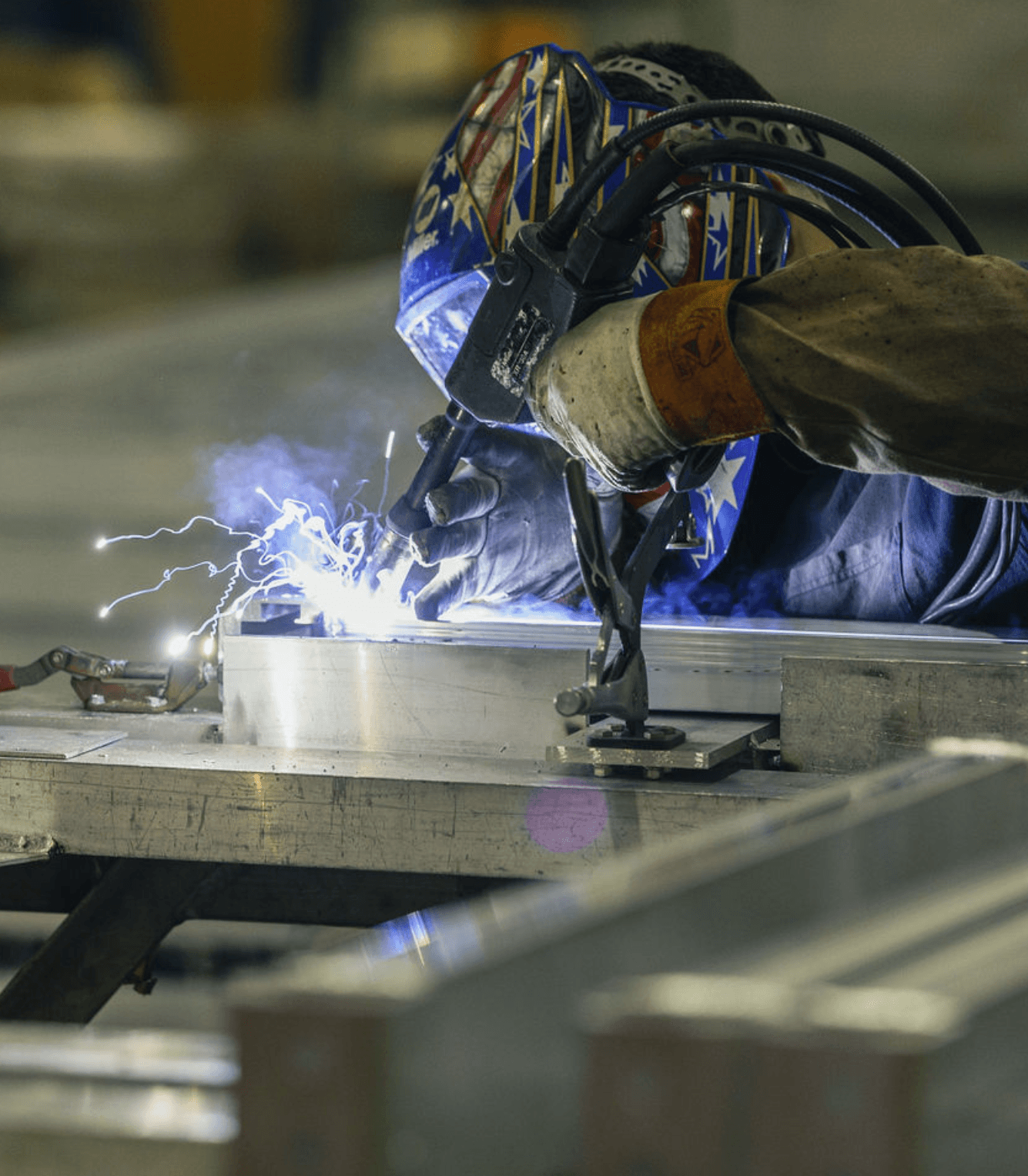 Rugged welder in a helmet and gloves welding a structural aluminum trailer frame on a fixture table