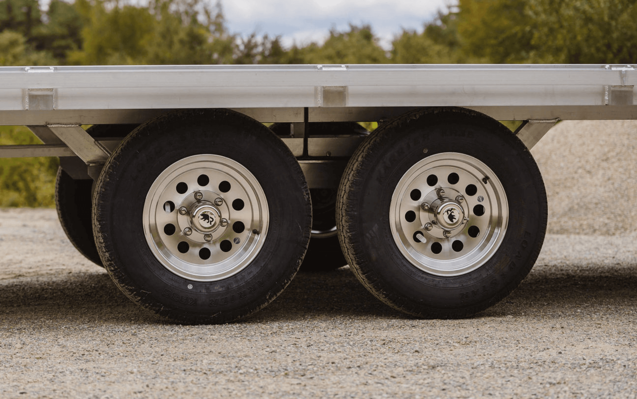 Ground-level view of tandem aluminum mod wheels and welded aluminum frame beneath a Rugged trailer deck.