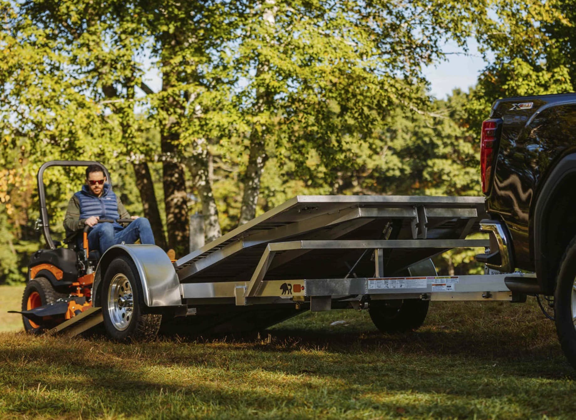 A Rugged tilt trailer with its deck tilted to grade behind a black pickup while a man drives a zero-turn mower off the ground end.