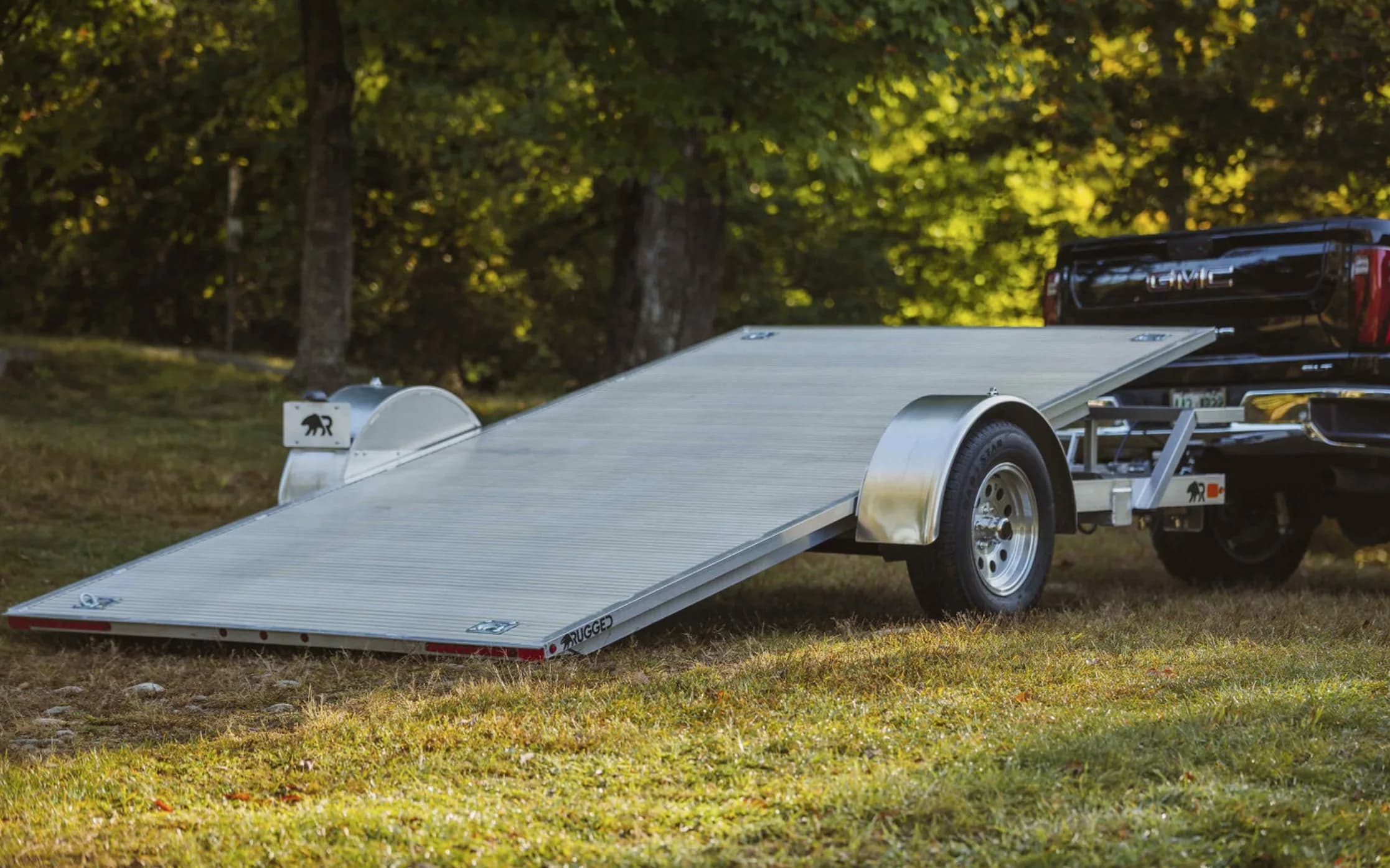A Rugged tilt trailer behind a pickup with its aluminum deck pivoted to the ground in loading position