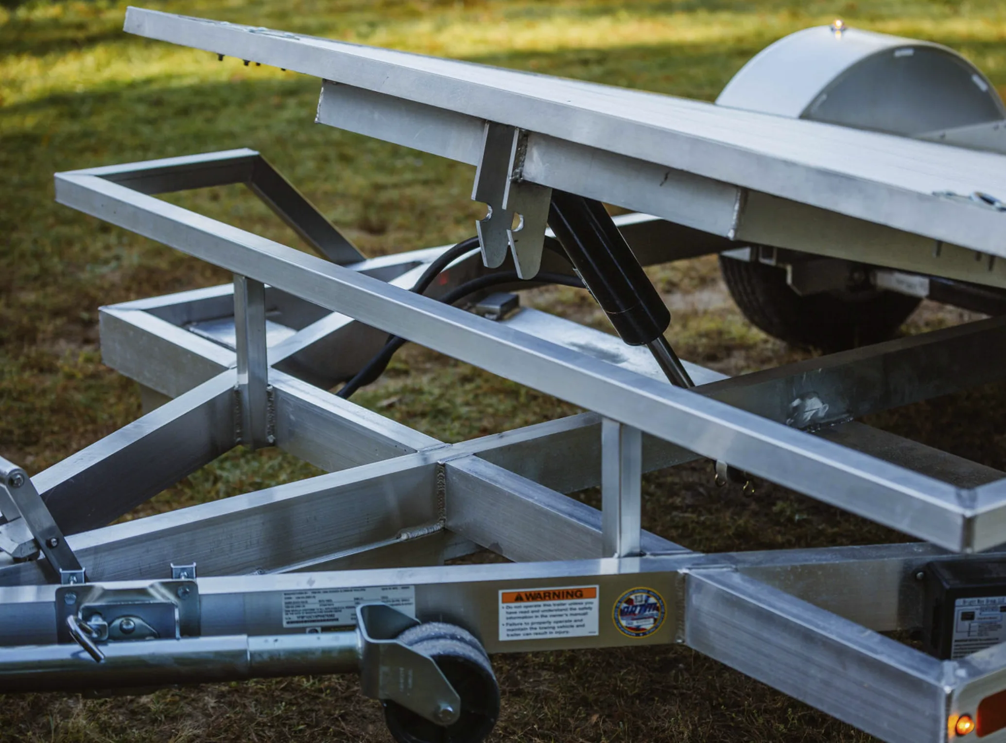 Close-up beneath a Rugged tilt trailer deck showing the TiltLock latch and hydraulic cylinder on the welded aluminum sub-frame