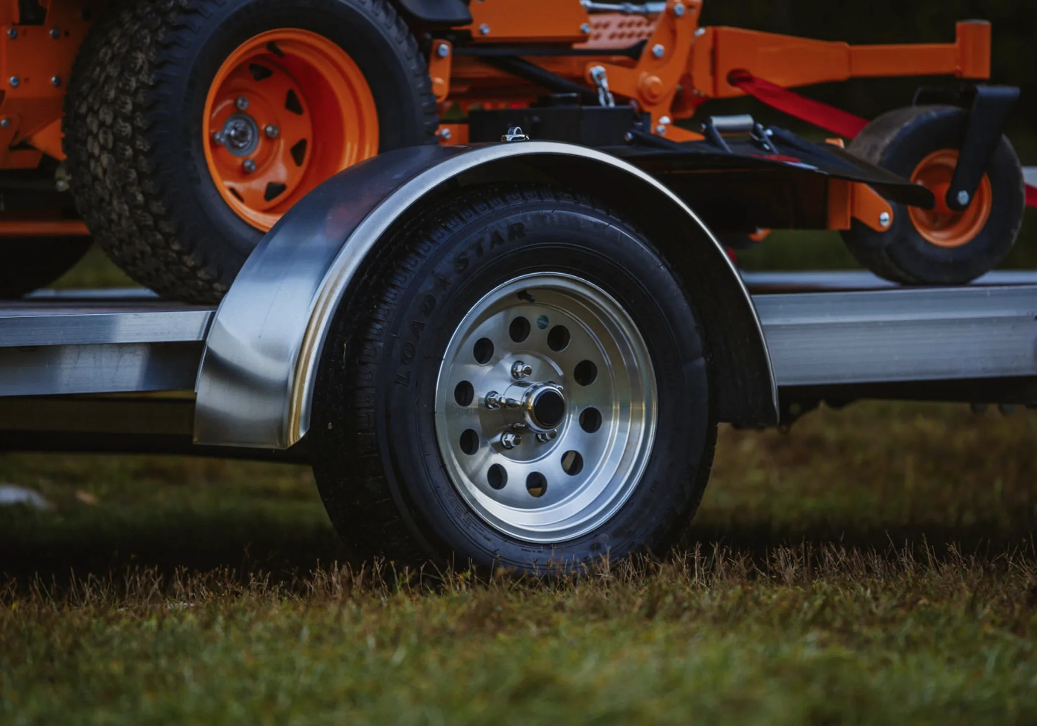 A 15-inch aluminum mod wheel under the fender of a Rugged tilt trailer carrying a zero-turn mower