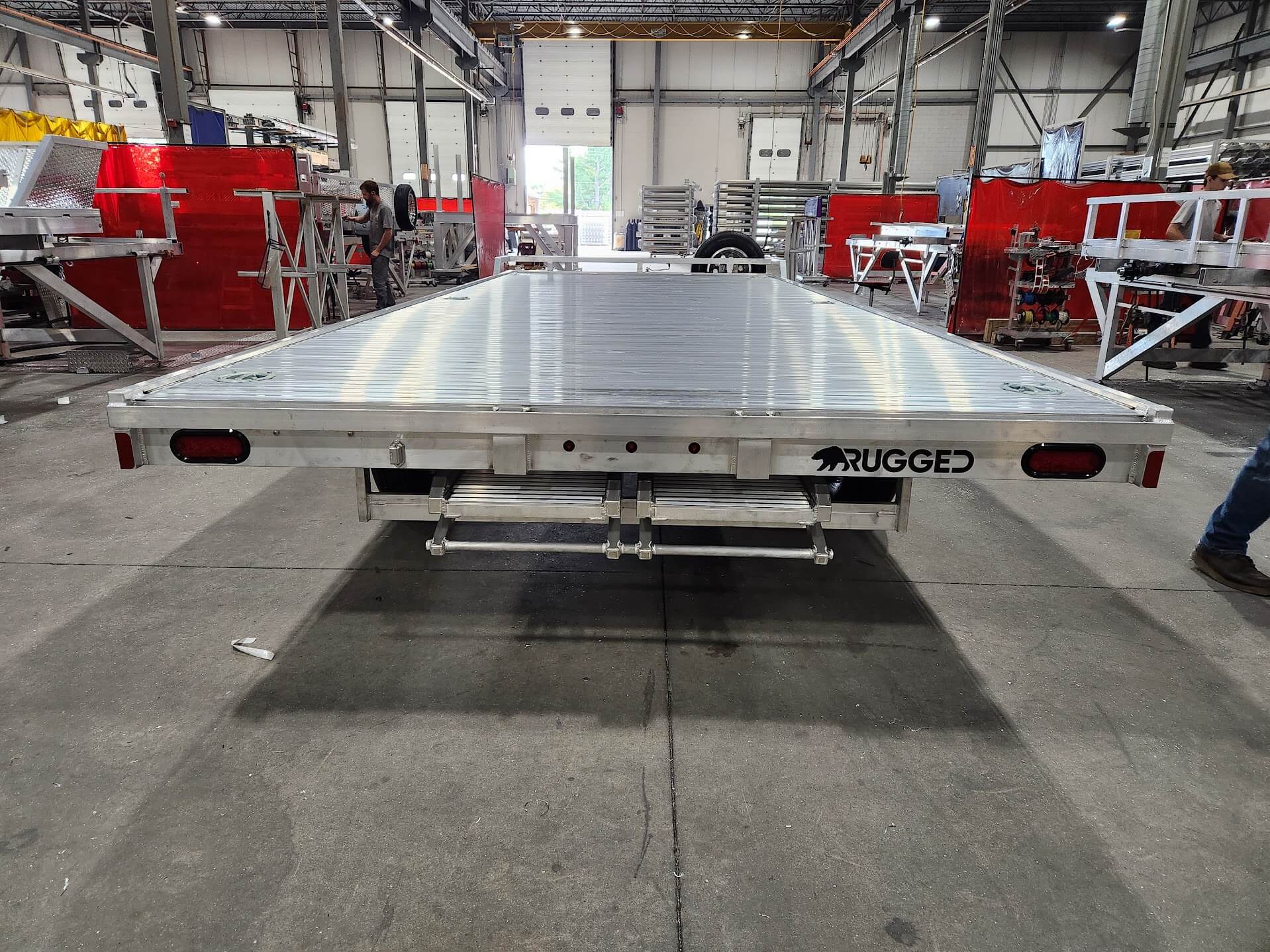 Rear view of a finished Rugged aluminum deckover trailer, showing the tail lights, rear beam and the Rugged wordmark, photographed on the floor before shipment