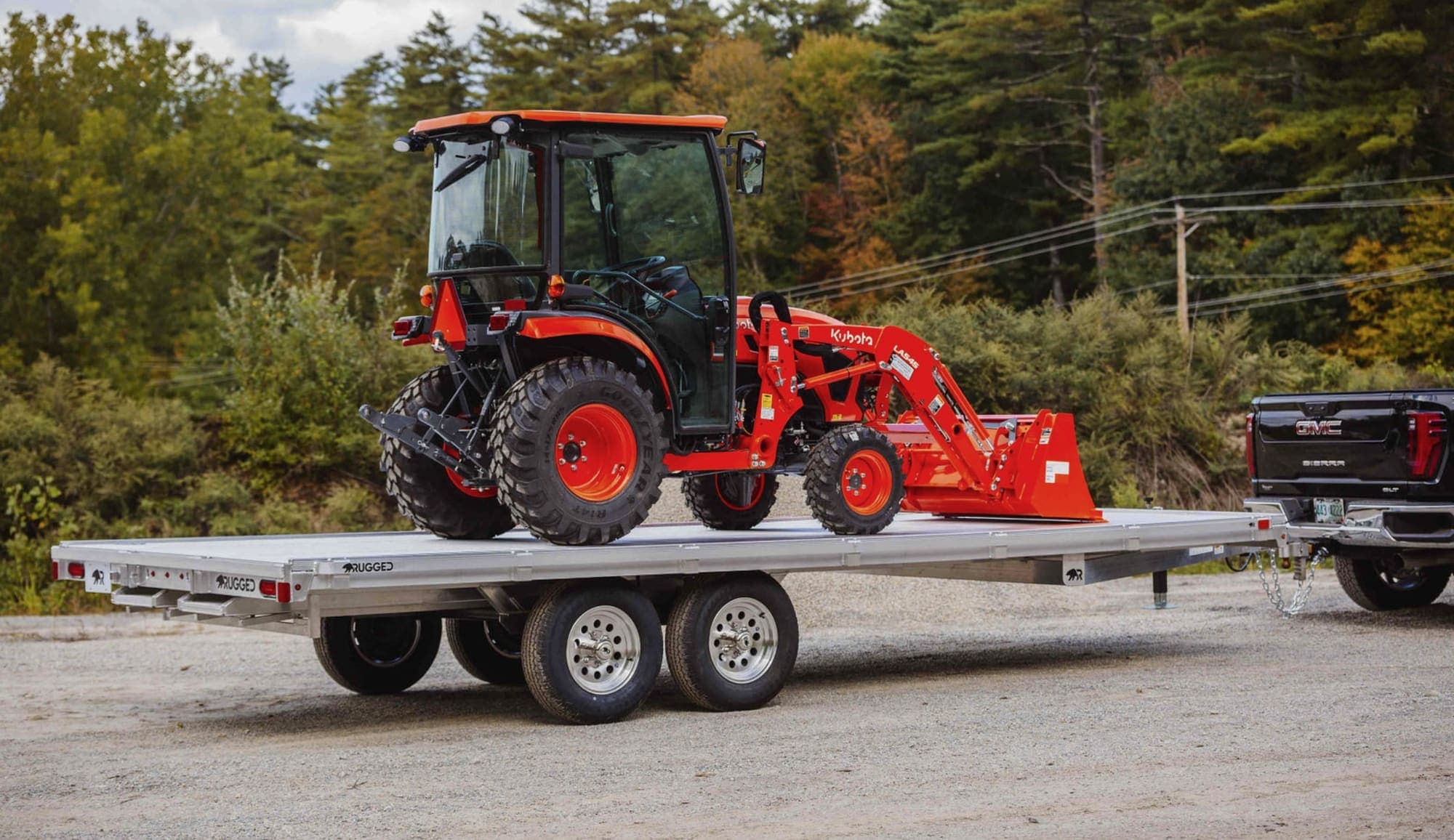 A red compact tractor with a front loader secured on a Rugged aluminum tandem-axle deckover trailer hitched to a black pickup truck.
