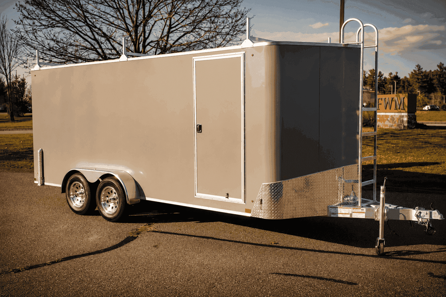 A gray tandem-axle enclosed cargo trailer with aluminum trim, a side access door, roof racks, and a front ladder in an outdoor lot.