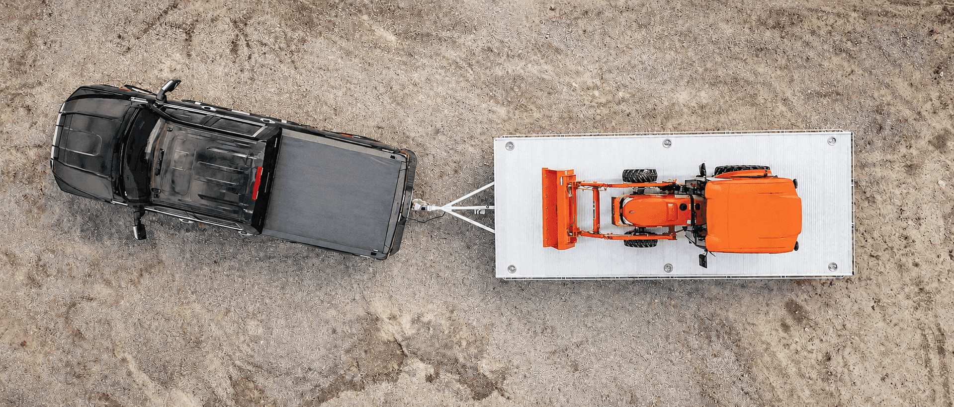 Top-down aerial view of a black pickup towing a Rugged aluminum deckover trailer carrying an orange compact tractor across a gravel yard