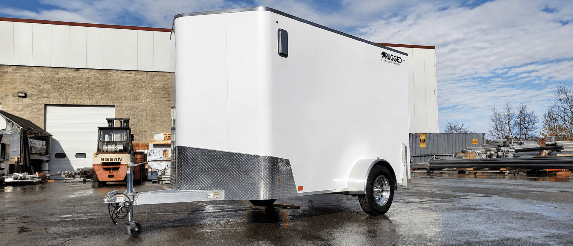 A white single-axle Rugged enclosed aluminum trailer with a diamond-plate rock guard parked in the factory yard, forklift and building behind