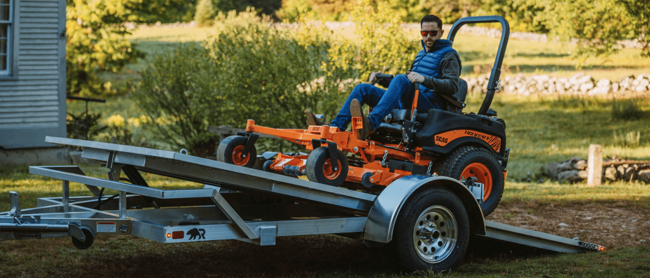 A man drives an orange zero-turn mower up the tilted deck of a Rugged single-axle aluminum tilt trailer, farmhouse and stone wall behind