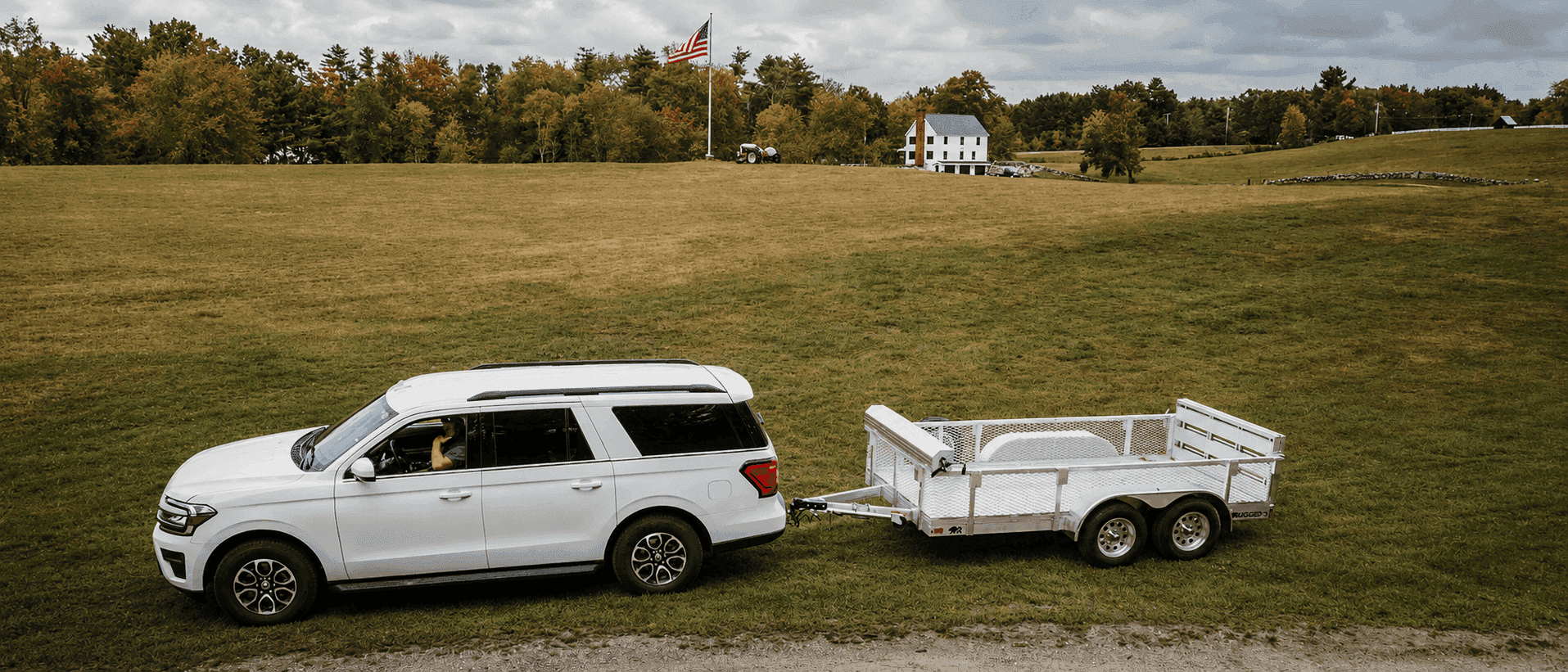 White SUV towing a Rugged tandem-axle open utility trailer with mesh side kit across a field, farmhouse and flag in the distance