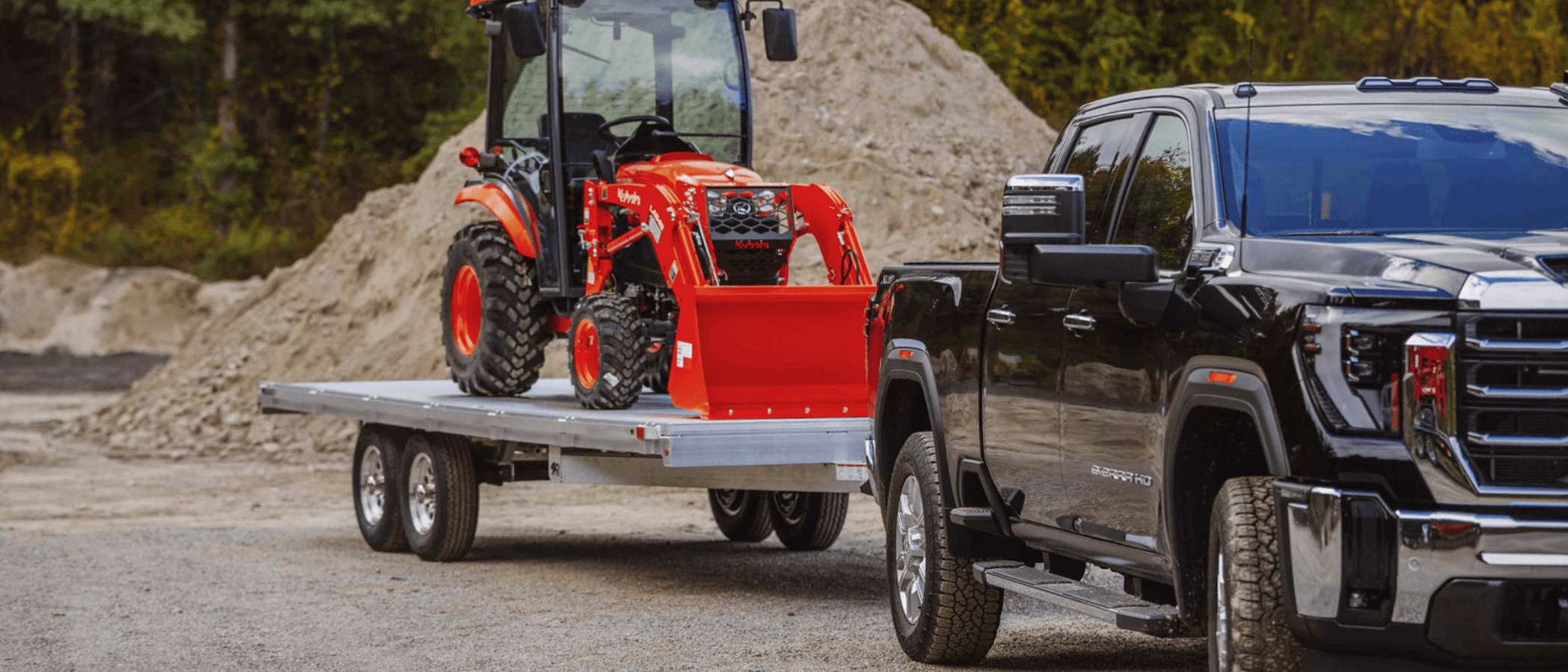 An orange compact tractor with a loader bucket rides on a Rugged tandem-axle aluminum deckover trailer towed by a black heavy-duty pickup past a gravel pile