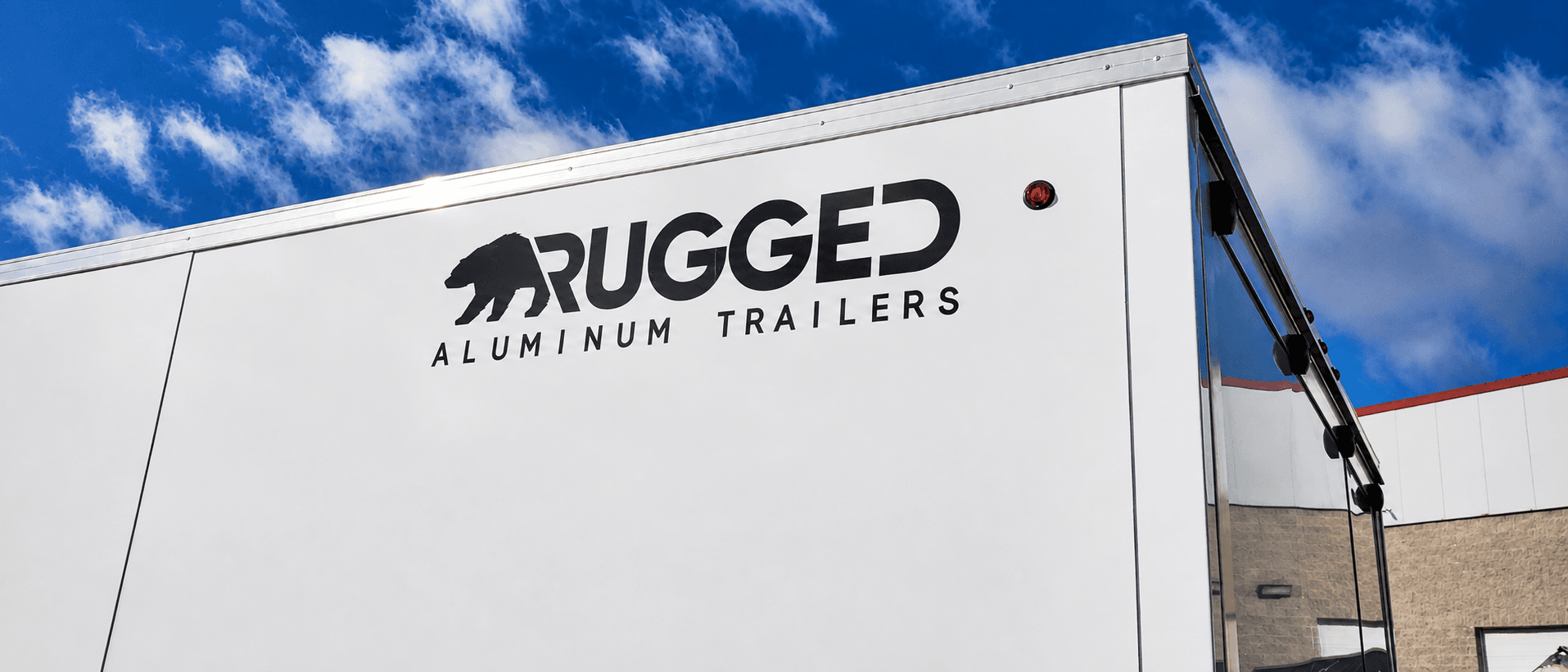 Low-angle view of a white Rugged enclosed trailer sidewall with the Rugged Aluminum Trailers wordmark and a red marker light against a blue sky