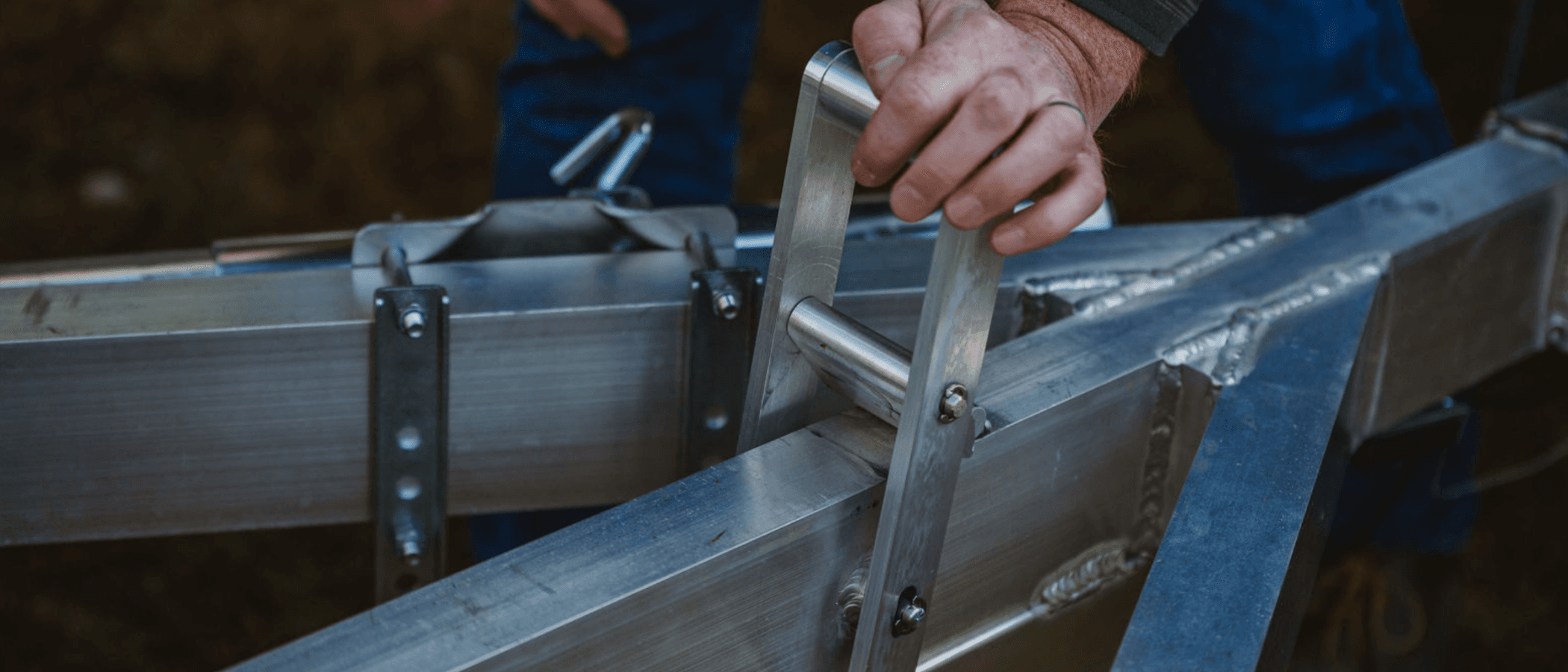 A hand grips the machined aluminum tilt lock lever on a Rugged tilt trailer frame, TIG weld beads visible on the members