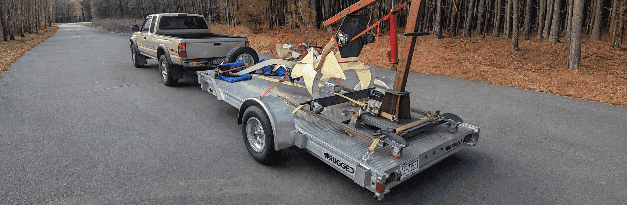 Rugged 6 × 12 utility trailer hauling an engine hoist and strapped cargo behind a Toyota Tacoma on a forest road