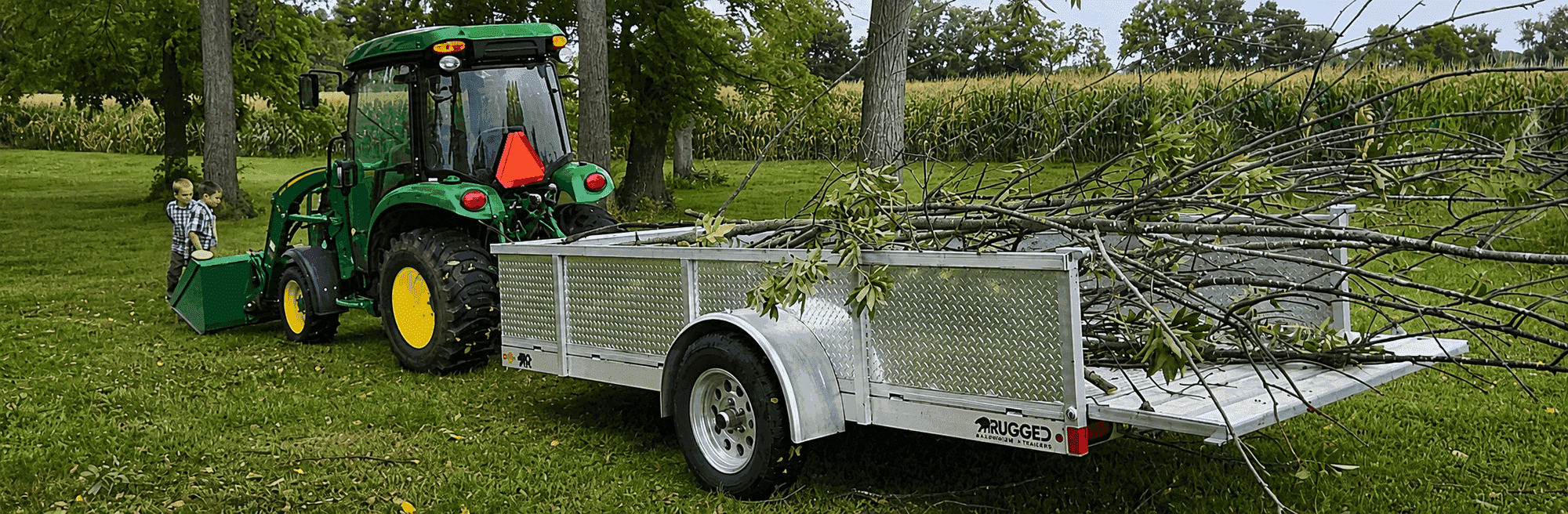 Rugged 6 × 14 utility trailer with diamond-plate side kit loaded with tree branches beside a John Deere tractor