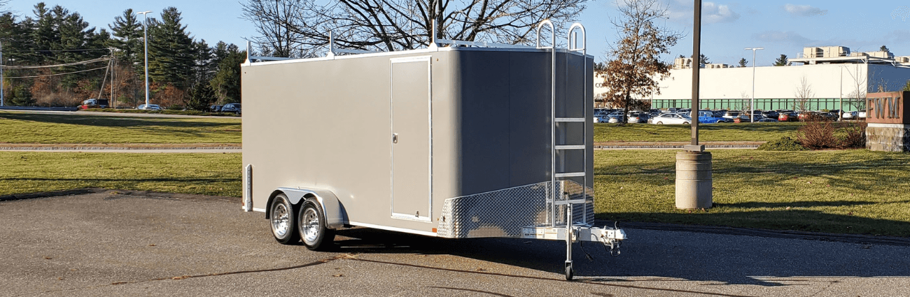 A gray tandem-axle Rugged enclosed trailer with a side man-door, diamond-plate rock guard, and roof rack with rear ladder, parked outside the FWM factory