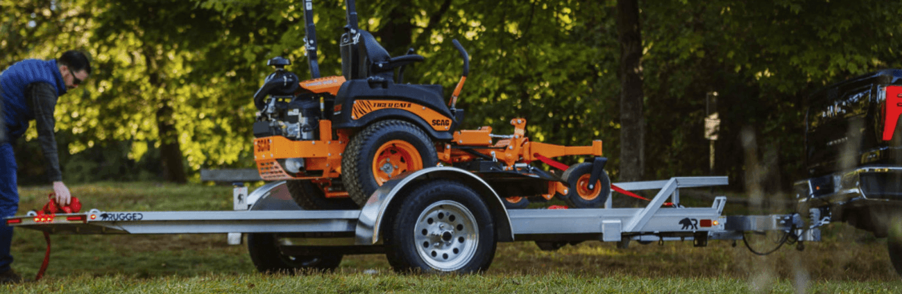 An orange zero-turn mower strapped down on a Rugged single-axle tilt trailer behind a black pickup while an owner secures the front strap