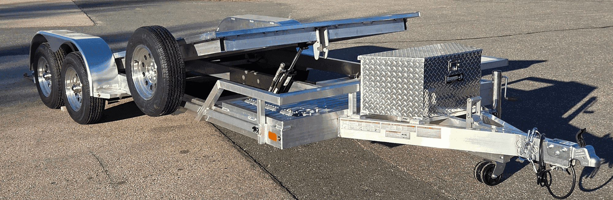 Rugged tandem-axle tilt trailer with the deck tilted up, hydraulic cylinder exposed and a diamond-plate tongue box on the frame