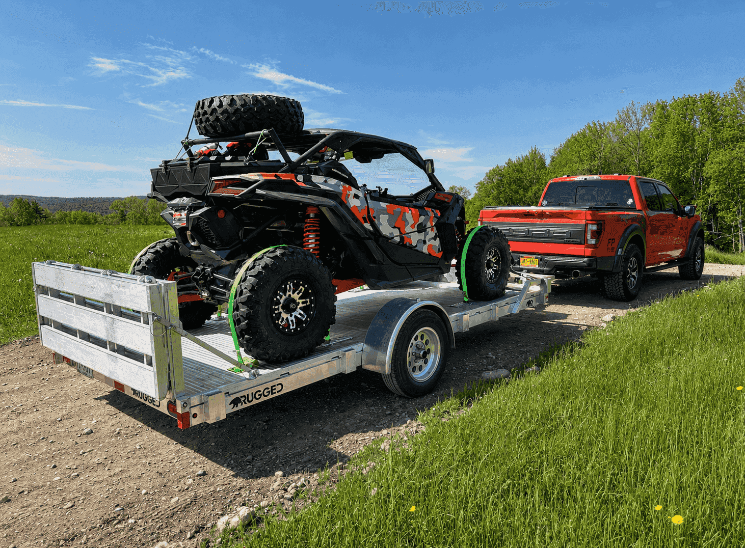 A camouflaged side-by-side secured to a Rugged all-aluminum utility trailer hitched to a red pickup on a sunny rural trail.