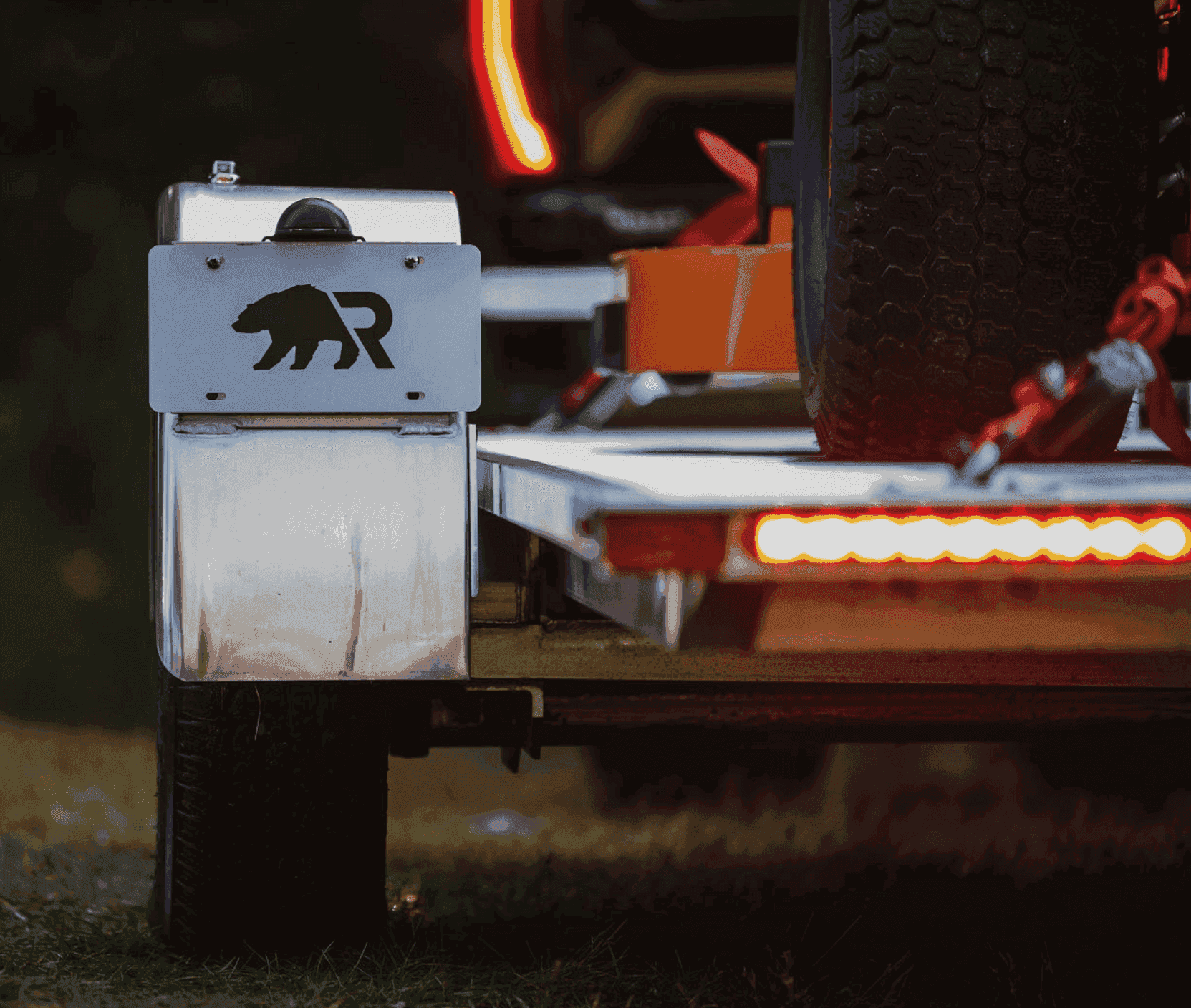 Rear detail of a Rugged aluminum trailer showing the branded metal box, spare tire, tie-down, and illuminated tail lights.