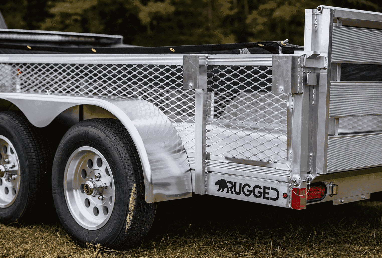 Rear quarter view of a Rugged all-aluminum utility trailer with mesh sides, tandem axles, and the Rugged wordmark on the fender skirt