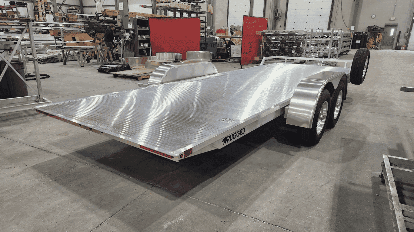 A Rugged aluminum tilt trailer on the shop floor, showing the diamond-plate deck, tandem axles, and the Rugged wordmark on the side rail