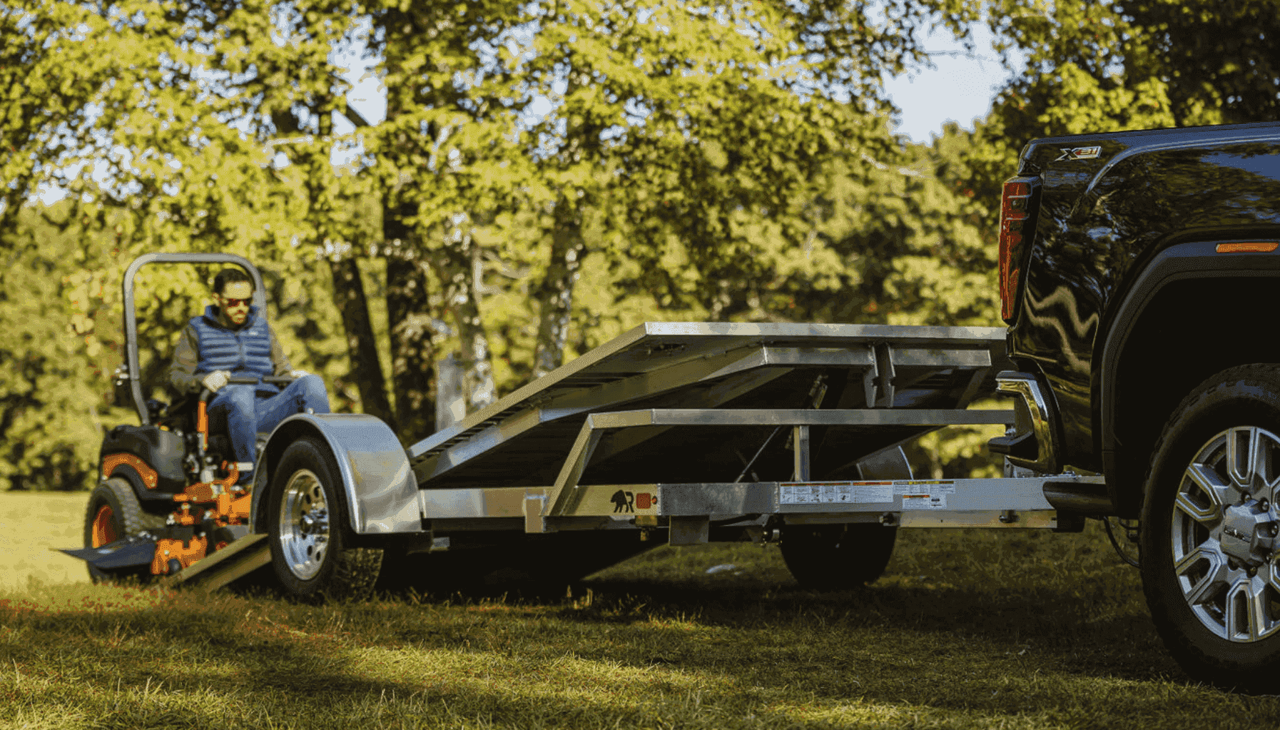 A customer drives an orange zero-turn mower onto a Rugged aluminum tilt trailer lowered to the grass behind a black pickup truck.