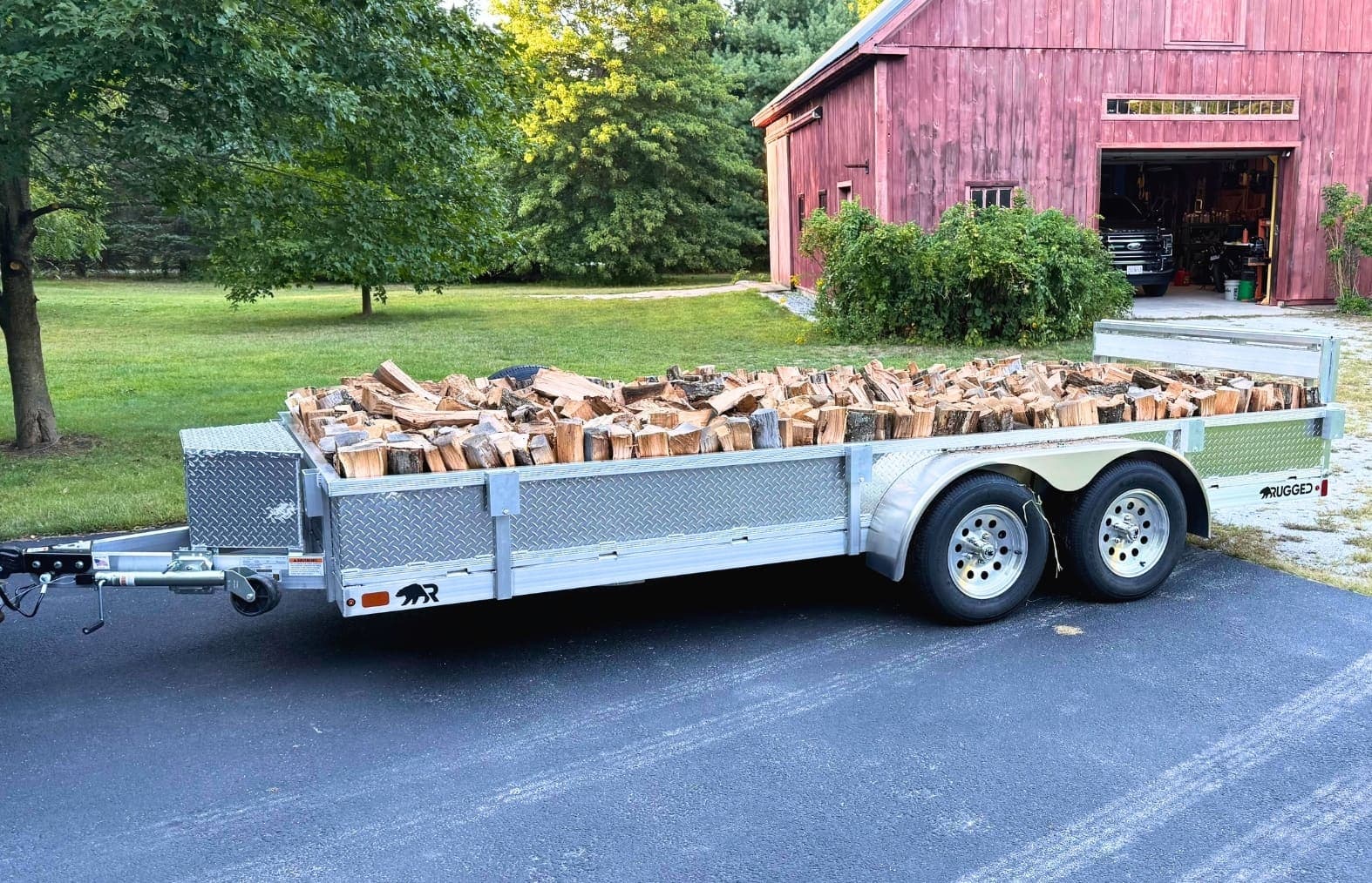 A Rugged tandem-axle all-aluminum utility trailer with diamond-plate sides, loaded with split firewood and parked on a driveway in front of a red New England barn.