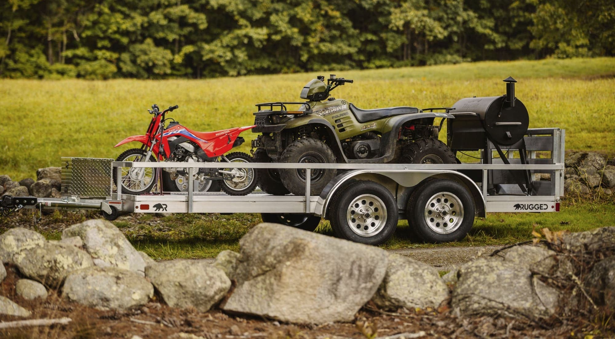A Rugged tandem-axle aluminum utility trailer loaded with a red dirt bike, an ATV, and an outdoor smoker in a grassy field.