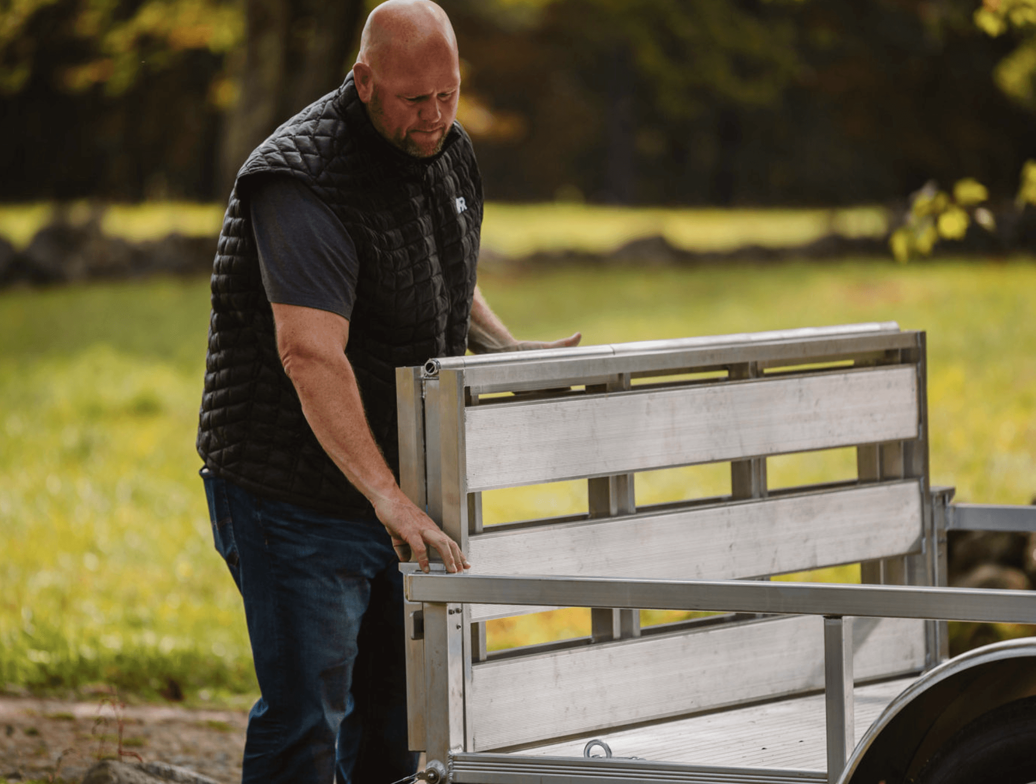 An owner folding down the aluminum tailgate of a Rugged utility trailer in a field.