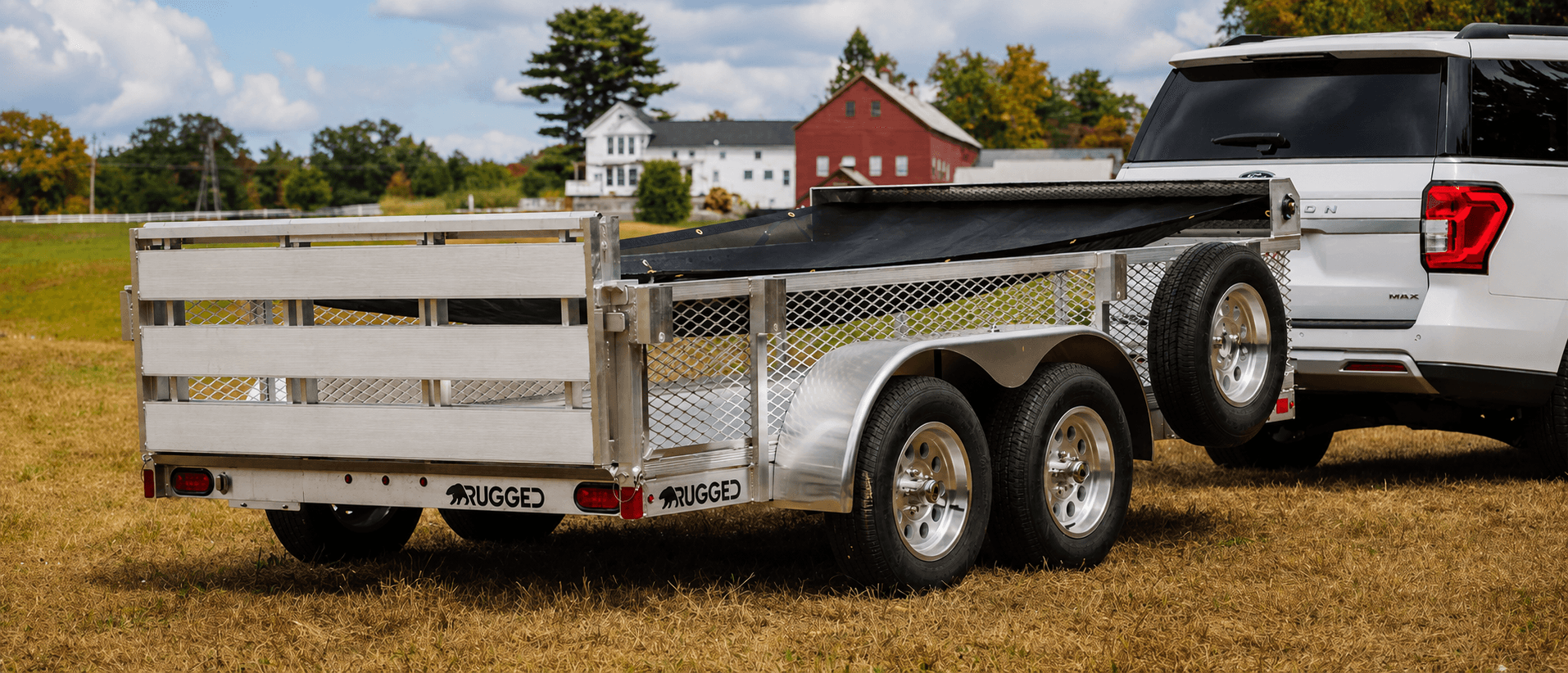 Rugged tandem-axle aluminum utility trailer with mesh sides and a black mesh tarp, hitched to a white Ford Expedition in front of a New England farm