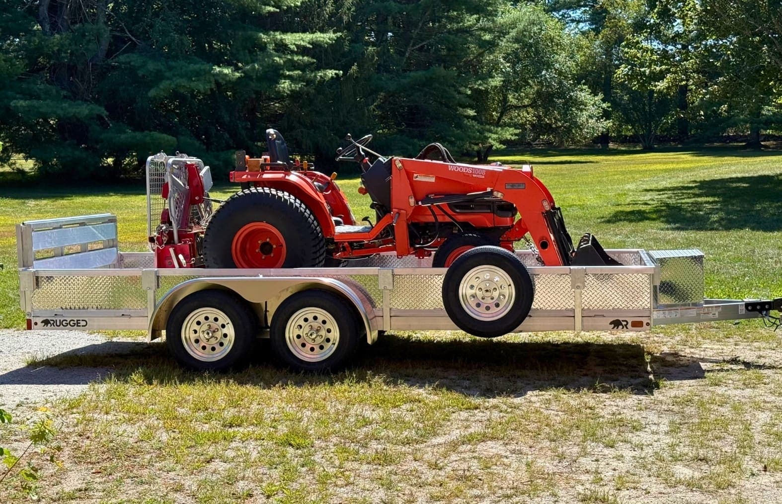 A Rugged tandem-axle all-aluminum utility trailer with diamond-plate sides hauling a small orange compact tractor, parked on a green lawn.